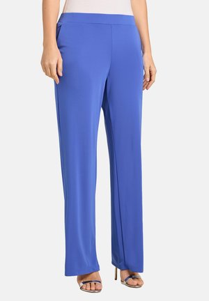 Kobieta w jaskrawo niebieskich szerokich spodniach z wysokim stanem oraz srebrnych sandałach na wysokim obcasie z odkrytymi palcami, stojąca na białym tle.