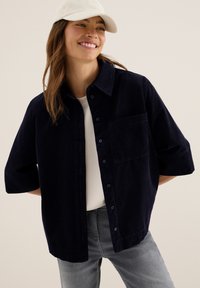 Lachende vrouw in een witte pet, donkerblauwe button-up ribfluweelshirt over een witte top, en grijze jeans, staande tegen een neutrale achtergrond.