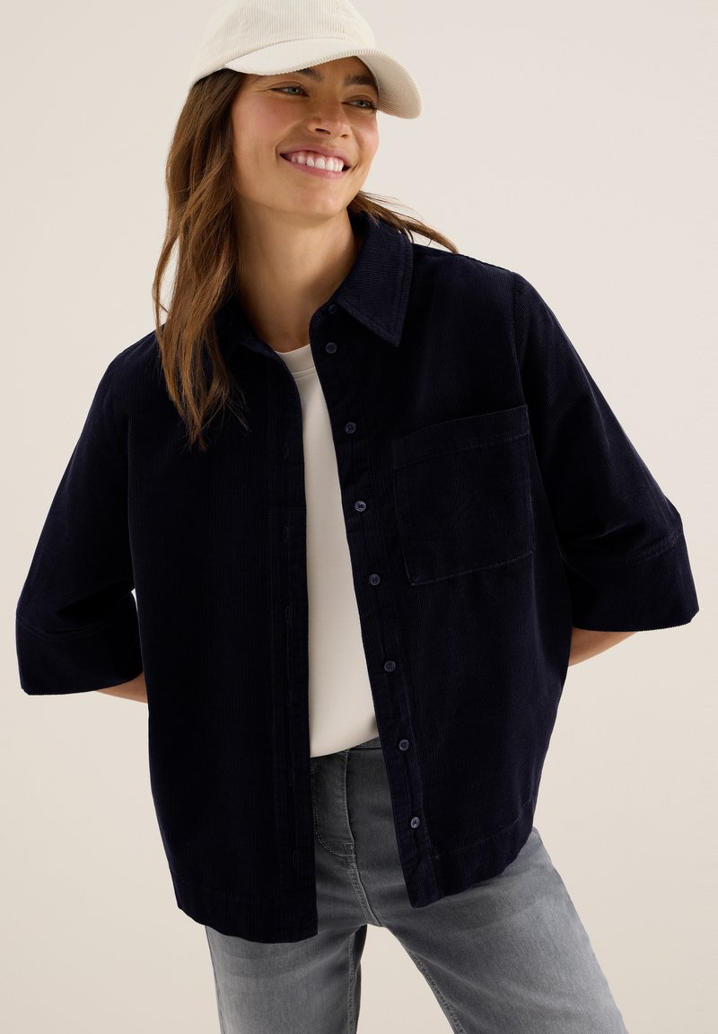 Lachende vrouw in een witte pet, donkerblauwe button-up ribfluweelshirt over een witte top, en grijze jeans, staande tegen een neutrale achtergrond.