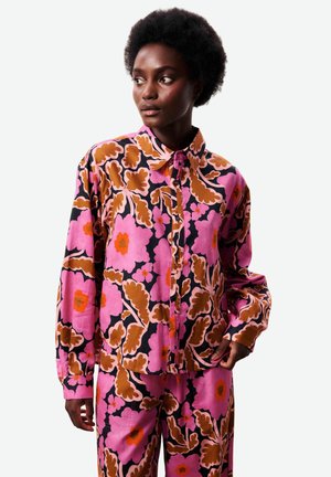 Femme portant une chemise boutonnée et un pantalon assortis à motif floral rose, marron et noir, regardant sur le côté avec une main dans la poche.
