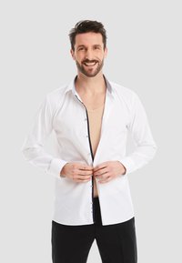 White shirt with a contrasting navy button placket and collar, long sleeves, and a tailored fit, paired with a beige undershirt.