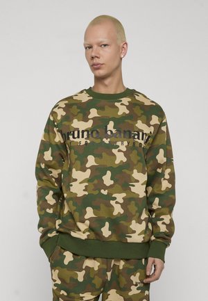 Osoba ubrana w zieloną, brązową i beżową bluzę oraz spodnie moro z napisem "bruno banani" na bluzie, stojąca z rękami w kieszeniach.