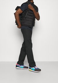 Schwarze Steppweste über einem dunkelblauen, gemusterten T-Shirt, schwarze Hose und mehrfarbige Sneakers mit blauen, pinken und schwarzen Akzenten.