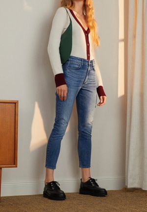 Frau trägt eine weiße Strickjacke mit bordeauxrotem Rand, blaue hoch taillierte Jeans, schwarze Schnürschuhe und eine grüne Schultertasche drinnen.
