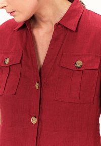 Rotes Leinenhemd mit Kragen, zwei Brusttaschen und beigen Knöpfen mit roten Nähten. Weiche Textur und lässiges Design.