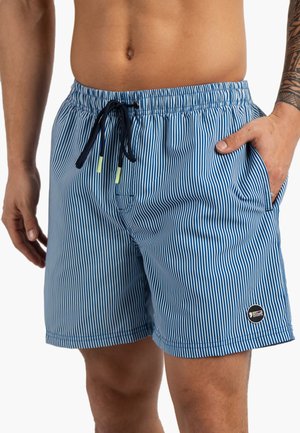 Torse masculin portant un short de bain à rayures verticales bleues et blanches avec un cordon noir et la main dans la poche, tatouage visible sur le bras.