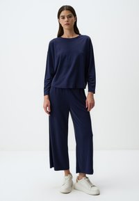 Navy Loungewear-Set aus weichem Stoff, bestehend aus einem locker sitzenden Langarmoberteil und weit geschnittenen Hosen, ergänzt durch weiße Sneakers.