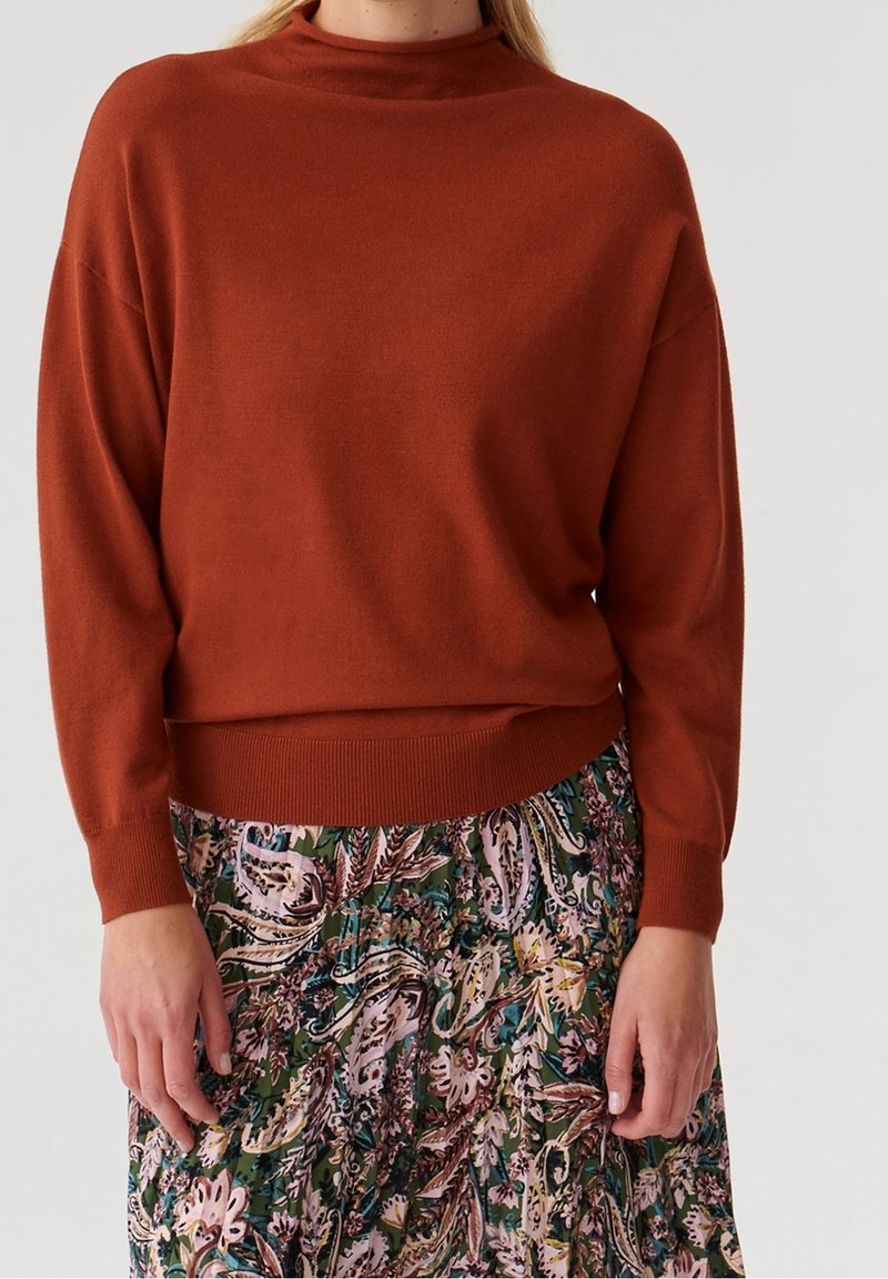 Femme portant un pull à manches longues couleur rouille, associé à une jupe taille haute à motifs floraux dans des tons verts, roses et beige.