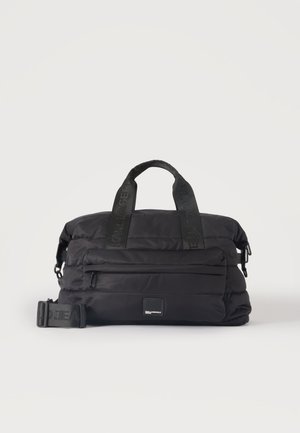 Sac de voyage noir matelassé avec deux poignées, une poche avant zippée et une sangle latérale réglable sur un fond uni.