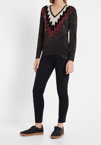 Pull noir à manches longues avec un col en V, présentant un motif zigzag multicolore en rouge, crème et noir, porté avec un jean noir et des baskets.