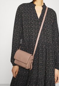 Křížová taška z měkké růžové kůže, obdélníkového tvaru, s klopou a nastavitelným popruhem, spárovaná s vzorovanými černými šaty.