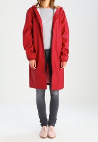 Czerwona wodoodporna peleryna przeciwdeszczowa z kapturem, z przodu z zamkiem błyskawicznym i elastycznymi mankietami, noszona na szarym topie i szarych dżinsach, zestawiona z jasnymi butami.