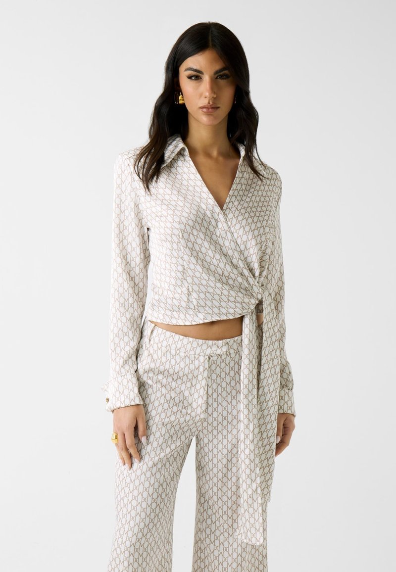 Mujer con cabello largo y oscuro que lleva una blusa cruzada blanca con estampado y pantalones anchos a juego sobre un fondo claro y liso.