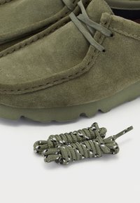 Mocassins en suédine vert olive avec des lacets assortis, présentant une texture douce et des coutures visibles, posés sur une semelle en caoutchouc texturée.