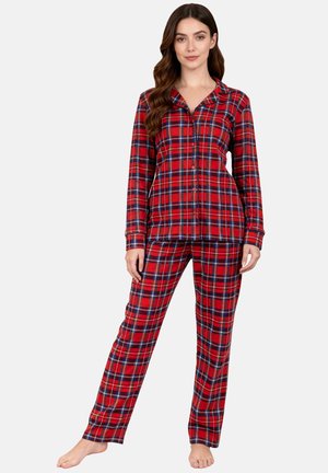 Kvinde står barfodet iført rød, blå og hvid ternet skjortebluse med lange ærmer og matchende lange pyjamasbukser på hvid baggrund.