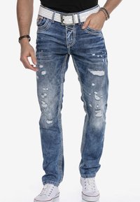 Blaue, distressed Jeans mit Slim-Fit, die große Risse, leichte Verwaschungen und kontrastierende Nähte an den Seiten aufweisen, kombiniert mit einem weißen, mit Nieten besetzten Gürtel.