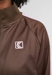 Brunotas užtrauktukas švarkas su tinkliniu tekstūru, turintis aukštą apykaklę ir šviesiai rožinius akcentus. Siuvinėtas logotipas kampelyje.