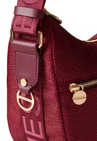 Burgundy textured shoulder bag with wide adjustable strap, gold zipper, and round gold zipper pull engraved with brand name.