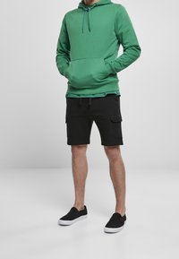 Grüner Hoodie mit einer Vordertasche, kombiniert mit schwarzen Cargo-Shorts mit zwei Seitentaschen. Dazu schwarze Slip-On-Schuhe mit weißen Sohlen.