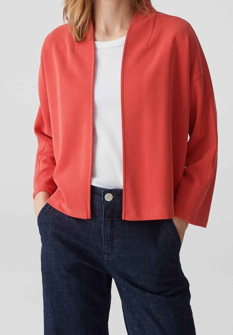 Femme portant une veste corail ample à ouverture devant par-dessus un top blanc côtelé et un jean bleu foncé taille haute, mains dans les poches.