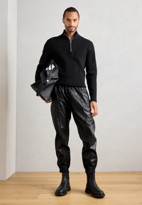 Hombre vistiendo un suéter negro de media cremallera, pantalones jogger de cuero negro, botas negras y sosteniendo una mochila de cuero negra contra una pared gris claro.