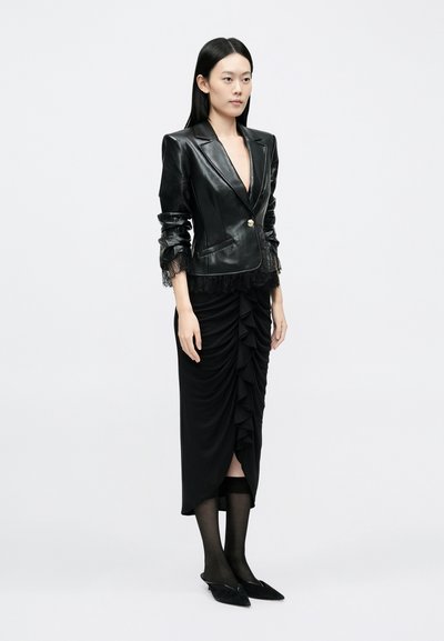 Fekete bőr blézer csipkés mandzsettával és fodros fekete szoknya. A modell áttetsző fekete harisnyát és hegyes orrú balerinacipőt visel. Egyszerű, testhezálló kialakítás.