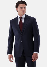 Marineblauer, maßgeschneiderter Anzug mit zweiknöpfiger Vorderseite, Spitzenrevers und zwei Vordertaschen. Dazu ein weißes Hemd und eine lachsrote Krawatte.