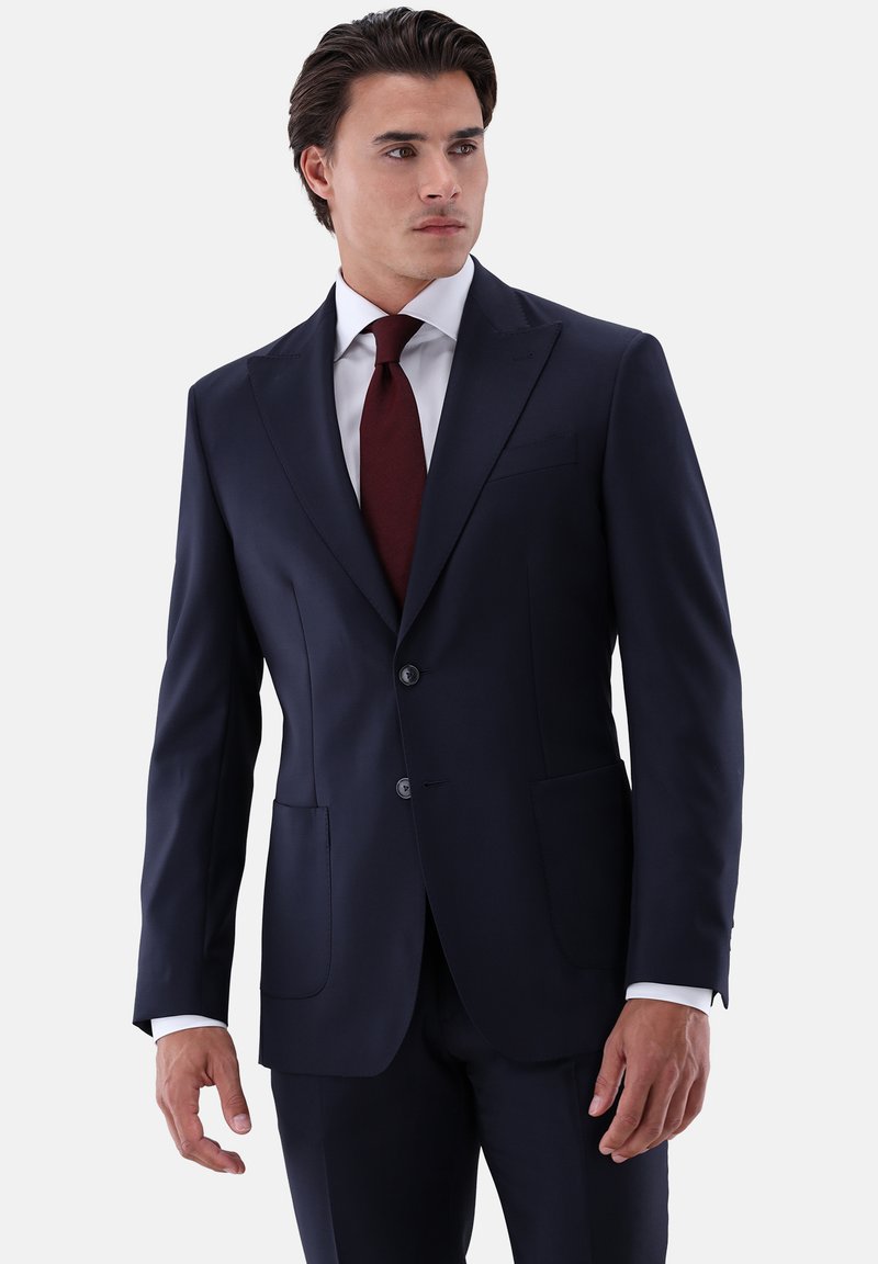 Marineblauer, maßgeschneiderter Anzug mit zweiknöpfiger Vorderseite, Spitzenrevers und zwei Vordertaschen. Dazu ein weißes Hemd und eine lachsrote Krawatte.