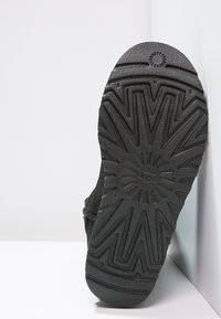Semelle de botte en caoutchouc noir avec motif en rayons de soleil, montrant le logo "UGG" au centre, photographiée sur fond blanc.