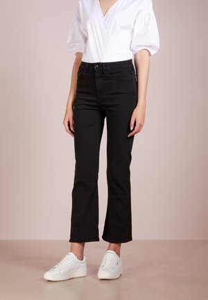 Frau trägt weiße Bluse mit Puffärmeln, hoch taillierte schwarze Jeans und weiße Plateau-Sneaker, steht auf einem beigen Boden vor einer beigen Wand.