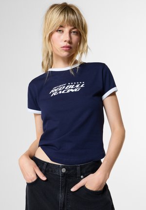 Русокоса жена с права коса, носеща тъмносиня тениска Red Bull Racing с бели кантове, стои с ръце в джобовете на черни дънки.