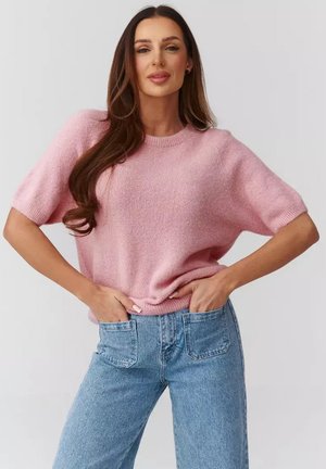 Kobieta z długimi brązowymi włosami, ubrana w jasnoróżowy sweter z krótkim rękawem i niebieskie dżinsy, z rękami w przednich kieszeniach.