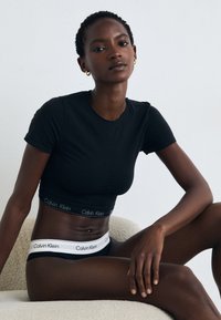 T-shirt nera aderente con maniche corte, abbinata a biancheria intima nera con una fascia in vita bianca con il marchio Calvin Klein. Materiale liscio e elastico.