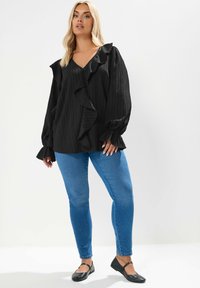 Blusa nera a maniche lunghe con costine verticali e dettagli di volant lungo il davanti e i polsini. Abbinata a jeans skinny blu e ballerine nere.
