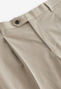 Beige Faltenhose aus strukturiertem Stoff, mit einem schwarzen Knopf und klassischem Bund. Der maßgeschneiderte Schnitt betont die Gesamtform.