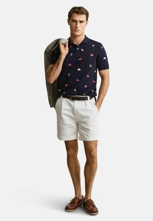 Man wearing navy polo with red crab and shell prints, white shorts, brown belt, and brown shoes, holding gray jacket over shoulder.