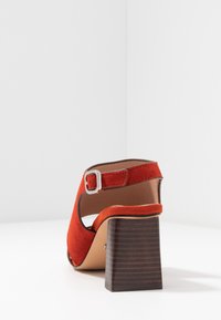 Červené semišové sandály na vysokém podpatku s kulatou špičkou a páskem přes kotník, s čtvercovým dřevěným podpatkem a stříbrnou sponou jako akcentem.