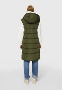 Chaleco puffer verde oliva con capucha, dividido por costuras horizontales, superpuesto a un suéter blanco, combinado con jeans azules remangados y zapatos beige.