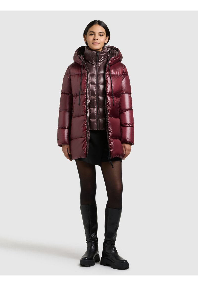 Moteris dėvi blizgią bordinę pūstinę striukę su kapišonu, juodą sijoną, permatomas pėdkelnes ir juodus kelių aukščio batus, stovinti prieš neutralią foną.