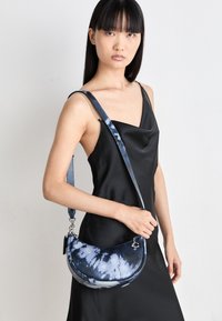 Blue tie-dye crossbody bag with a curved shape, silver hardware, and a strap featuring a chain accent, set against a black dress.