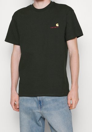 Osoba ubrana w czarną koszulkę z logo Carhartt i jasnoniebieskie dżinsy, stojąca z luźno opuszczonymi ramionami na jednolitym białym tle.