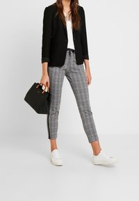 Blazer nero, top bianco, pantaloni grigi a scacchi con strisce laterali, borsa nera con manici marroni e sneakers bianche.