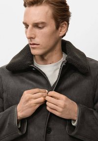 Giacca in suede grigia con colletto in shearling e dettagli in pelle. Il modello sta allacciando la giacca. Lo sfondo è bianco uniforme.