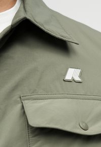 Κοντινό πλάνο ανοιχτόχρωμου ελιάς πράσινου μπουφάν με τσέπη με καπάκι και λευκό-γκρι γεωμετρικό λογότυπο στο στήθος.