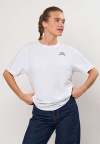 Femme aux cheveux tressés portant un t-shirt blanc avec un logo et un jean bleu foncé, debout les mains sur les hanches, regardant sur le côté.