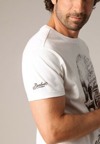 T-shirt en coton blanc avec un imprimé graphique de crâne sur le devant et "Deelux" brodé en marron sur la manche droite. Manches courtes.