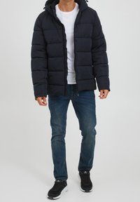 Dunkelblauer Puffer-Jacke mit Kapuze, gestepptem Design und Frontreißverschluss. Getragen über einem weißen T-Shirt, kombiniert mit blauen Jeans und schwarzen Sneakers.
