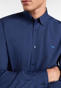 Temno modra srajca s kratkimi rokavi in klasičnim ovratnikom, ki na prsih vsebuje subtilen svetlo moder logotip psa in usklajene gumbe. Gladka tekstura.