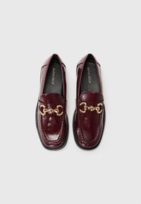 Mocassins en cuir verni bordeaux avec un accent en chaîne dorée, présentant un bout arrondi et une surface texturée, montés sur une semelle noire.