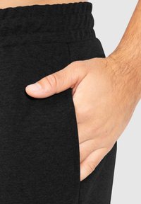 Schwarze Sporthosen aus strukturiertem Stoff, mit elastischem Bund und Seitentasche, in der eine Hand ruht.